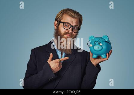 Close-up del barbuto Hipster imprenditore tenendo blue Piggybank con occhiali puntando ad esso con l'indice della mano isolato su sfondo blu. Posi Foto Stock