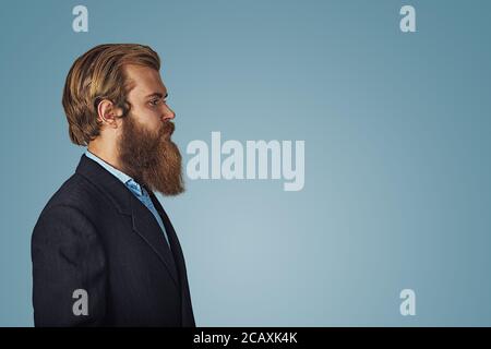 Vista laterale ritratto di grave alla moda hipster barbuto uomo bello in abito nero e camicia blu isolato su sfondo blu. Faccia negativa express Foto Stock