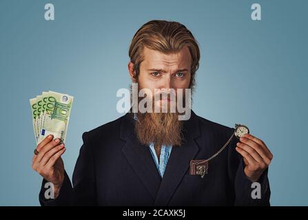 Il tempo è denaro concetto. Bello barbuto hipster grave imprenditore uomo con moneta contante e retrò orologio da tasca isolato su sfondo blu. Negativ Foto Stock