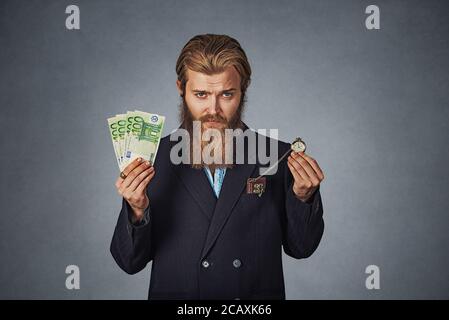 Il tempo è denaro concetto. Bello barbuto hipster imprenditore uomo con denaro contante in euro e in valuta retrò orologio da tasca isolato su sfondo grigio. Serie FR Foto Stock