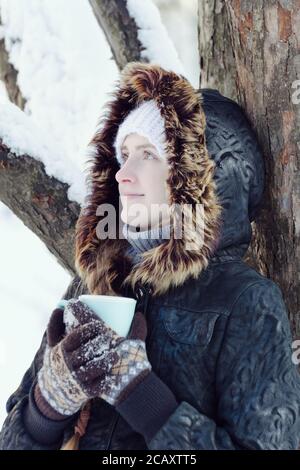 Ragazza in una giacca con cappuccio appoggiando un albero con una tazza di tè. Sfondo invernale Foto Stock