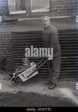 anni '50, storico, fuori in un cortile, accanto ad un capannone di metallo corrugato, un nonno che indossa tute, spingendo un carretto di legno a ruota singola, possibly un fatto in casa, dando la sua giovane nipote, che è seduto in esso, un giro. Foto Stock