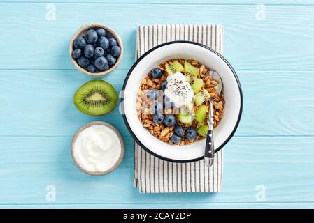 Granola al miele di avena con frutti di bosco, kiwi e yogurt greco su sfondo blu teal. Vista dall'alto piatto posate food. Concetto di dieta, mangiare pulito Foto Stock