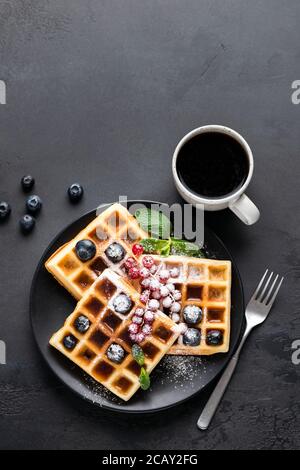 Waffle belgi quadrati con frutti di bosco e zucchero a velo e una tazza di caffè nero su fondo nero in cemento. Spazio di copia della vista superiore per testo o disegno Foto Stock