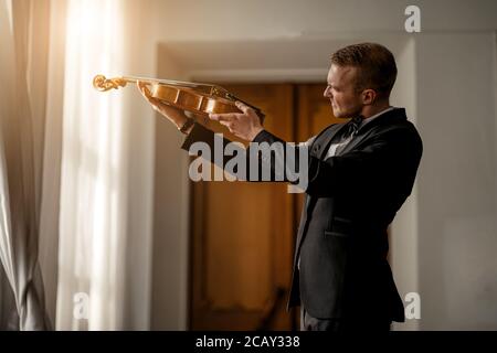 violinista caucasico maschio professionista di talento guarda il suo strumento, controllando le corde sul violino prima di eseguire la musica Foto Stock