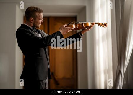 giovane violinista caucasico maschile professionista che studia e controlla le corde del violino prima del concerto. l'uomo tiene in mano il nuovo violino e lo guarda Foto Stock