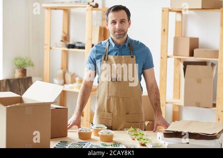 Vita in su ritratto di uomo bearded maturo che indossa grembiule e sorridente alla macchina fotografica in piedi da tavolo di legno con le parti individuali del cibo pronte per l'imballaggio, operaio nel servizio di consegna del cibo, spazio di copia Foto Stock