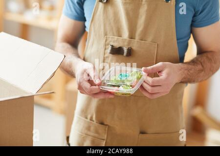 Primo piano di un uomo irriconoscibile che indossa grembiule imballaggio singole porzioni di cibo, lavoratore di servizio di consegna di cibo, spazio di copia Foto Stock