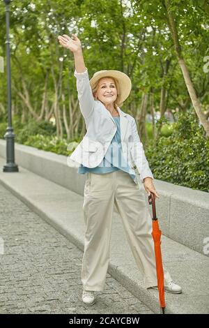 Anziana bianca bella signora di circa 62 anni sta viaggiando mentre in piedi sul marciapiede nel parco pubblico. È cordiale e allegra, vestita in cas Foto Stock