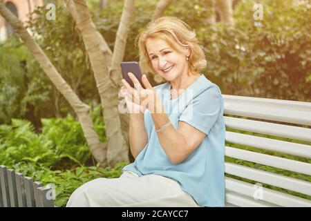 Bella donna bianca invecchiata circa 60 anni sta usando il suo smartphone mentre si siede sulla panchina nel parco pubblico. Sta sorridendo al suo dispositivo. Guarda Foto Stock