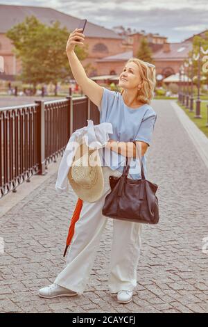 Allegra donna bianca caucasica invecchiata, di circa 60 anni, cammina nel parco pubblico e scatta foto con l'aiuto del suo smartphone. Ha casual Foto Stock