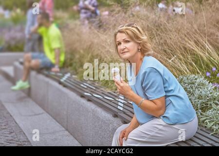 Anziano bianco attraente signora 62 anni è seduta sopra il banco con markup di divaricamento sociale nel parco pubblico durante epidemia di covid-19 e mangiare i Foto Stock