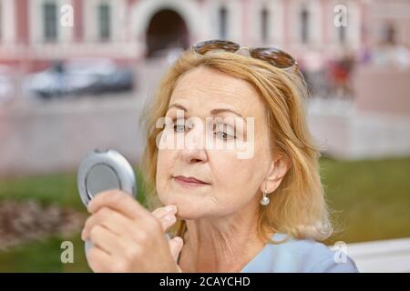 Invecchiato bianco caucasico bella donna d'affari circa 62 anni sta camminando nell'area del centro e guardando nello specchio. Indossa occhiali da sole e sembra più saggia Foto Stock