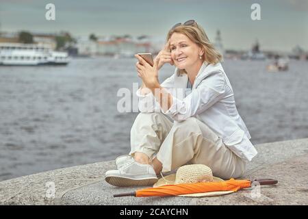 Anziana bianca bella donna sorridente 62 anni è seduta vicino al fiume mentre camminando nella zona del centro con il telefono delle cellule nelle mani. È attiva e. Foto Stock