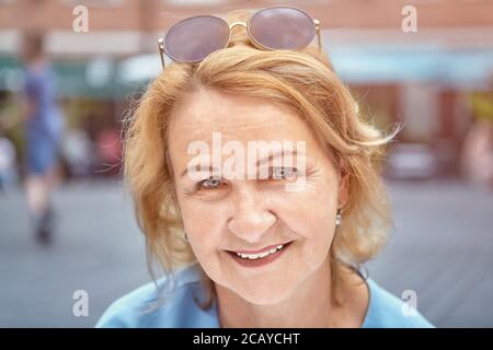 Ritratto di bella signora bianca invecchiata di circa 60 anni nel parco pubblico. Sorride e guarda attiva, affascinante e amichevole donna. Indossa occhiali da sole Foto Stock