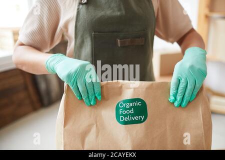 Primo piano di lavoratrice non riconoscibile che indossa guanti e tiene in mano un sacchetto di carta artigianale mentre gli ordini di imballaggio al servizio di consegna del cibo, spazio di copia Foto Stock
