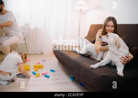 bella famiglia amichevole trascorrere il tempo libero a casa insieme, tutti sono impegnati con i propri interessi, madre giocare con cane animale domestico, figlia piccola w Foto Stock