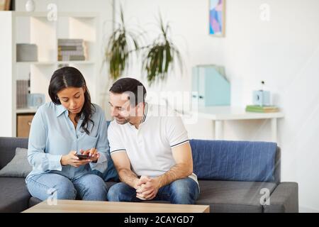 Ritratto di giovane donna di razza mista che mostra smartphone a un collega maschile mentre si siede sul divano in ufficio interno, copia spazio Foto Stock