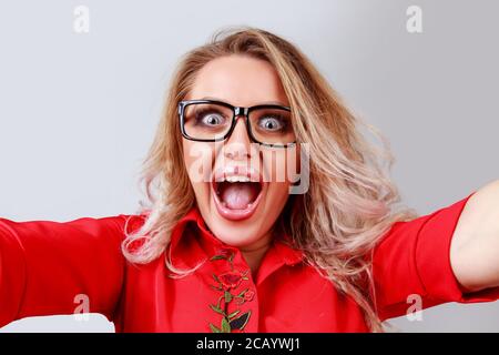Donna bionda eccitata in occhiali con gli occhi e la bocca spalancati si apre prendendo selfie e guardando la fotocamera su sfondo grigio. Foto Stock