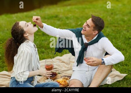 Ritratto di giovane coppia spensierata godendo di una data romantica all'aperto mentre si siede su erba verde vicino al lago e si mangia frutta Foto Stock