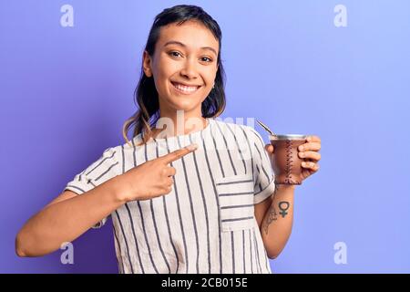 Giovane donna bevendo l'infusione di compagno sorridente felice indicando con mano e dito Foto Stock