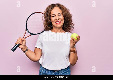 Media età bella sportivo che gioca a tennis tenendo racchetta e palla su sfondo bianco guardando positivo e felice in piedi e sorridendo con una c Foto Stock