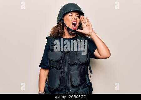 Bella donna soldato di mezza età che indossa uniforme antiproiettile e casco di sicurezza gridando e urlando forte a fianco con la mano sulla bocca. Comunicazioni Foto Stock