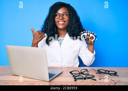 Bella donna africana che tiene occhiali da ottometria puntando il pollice verso il lato sorridendo felice con la bocca aperta Foto Stock