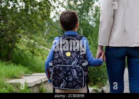 Il genitore e l'allievo della scuola primaria vanno di pari passo. Donna e ragazzo con zaino dietro la schiena. Foto Stock