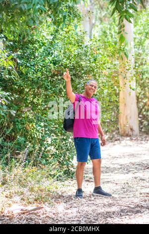 Ritratto di un uomo anziano positivo che porta con sé uno zaino, puntando con un dito indice, guardando la fotocamera e sorridendo. Fit vecchio su un viaggio escursionistico. Foto Stock