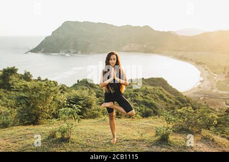 Giovane donna che pratica yoga posa al tramonto con splendida vista sull'oceano e sulle montagne. Sensibilità alla natura. Stile di vita attivo. Concetto spirituale ed emotivo. Foto Stock