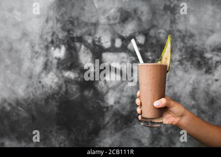 Bicchiere di latte fresco al cioccolato freddo agita in mani di donna sullo sfondo di cemento grigio parete testurizzata. Frullato di cacao con pezzo di banana come decorazione. Concetto estivo. Foto Stock