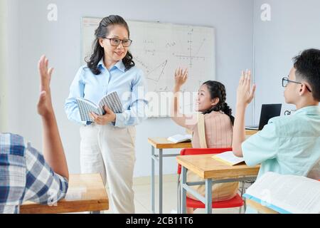 Sorridente insegnante di scienza matura che fa domande e guarda gli studenti alzando le mani e desiderando rispondere Foto Stock