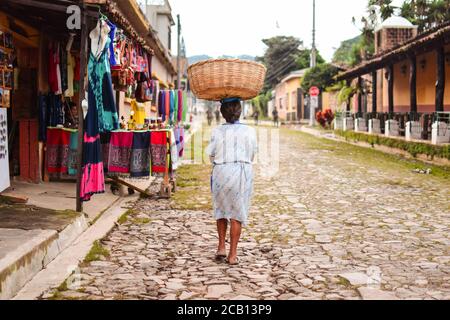 Concepcion de Ataco / El Salvador - 29 ottobre 2017: Donna Salvadorana che cammina lungo un sentiero di pietra con un cesto sulla testa e soldati pattugliando sullo sfondo Foto Stock