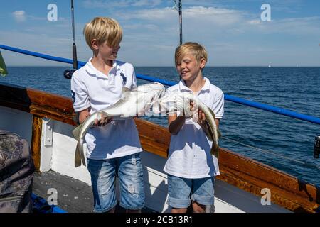 Malmo, Svezia - 6 agosto 2020: Due ragazzi in un viaggio di pesca con la famiglia mostrano la loro cattura fresca di merluzzo. Oceano blu e cielo sullo sfondo Foto Stock