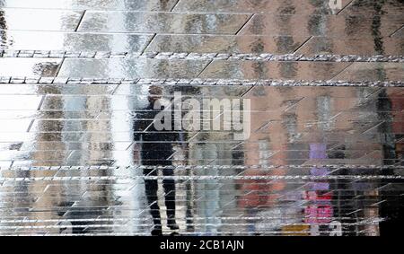 Sfocata riflessione ombra silhouette su bagnata puddle di un uomo che cammina una strada della città in una giornata piovosa, sfondo astratto Foto Stock