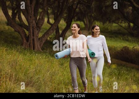 Due donne allegre che camminano in un parco con stuoie per lo yoga. Sorridente donne amici andare per un allenamento all'aperto. Foto Stock