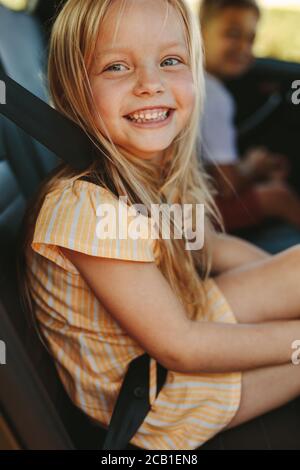 Primo piano di una ragazza carina seduto sul sedile posteriore di auto che indossa la cintura di sicurezza. Ragazza allegra che si diverse a viaggiare in auto. Foto Stock