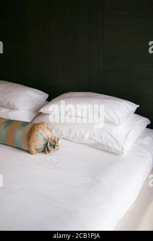 Un letto con una coperta bianca e cuscini di diverse dimensioni e un cuscino allungato in una camera con pareti scure. Foto Stock