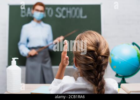 vista posteriore della studentessa con la mano sollevata, e insegnante con bastone di puntamento vicino alla lavagna Foto Stock