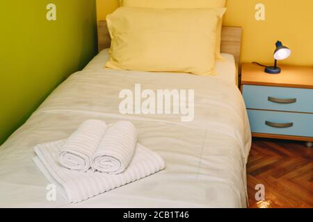 Un letto singolo con cuscini gialli e una coperta bianca in una camera con pareti gialle e verdi, un comodino di legno blu con una lampada da lettura e terry Foto Stock