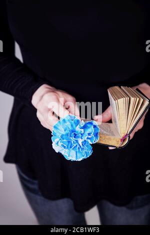 Mani femminili che tengono il vecchio libro e fiore blu di garofano su uno sfondo nero. Spazio di copia Foto Stock