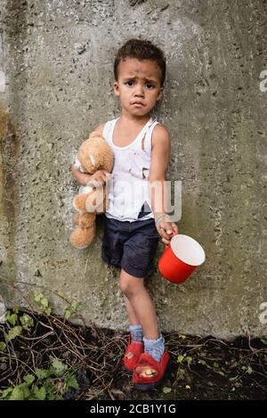 Insoddisfatto bambino afroamericano in vestiti sporchi che mendicante le elemosine vicino muro di cemento in baraccopoli Foto Stock