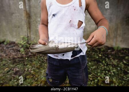 Vista ritagliata del povero ragazzo afro-americano che tiene una piastra di metallo e cucchiaio in baraccopoli Foto Stock