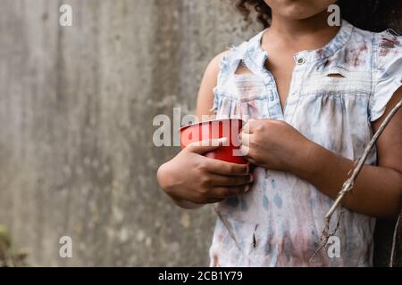 Vista tagliata del povero capretto afroamericano in vestiti sporchi tenendo una tazza di metallo su una strada urbana Foto Stock