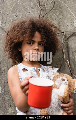 Fuoco selettivo del bambino afro-americano disgraziato con il teddy sporco orso elemosina mendicante su strada urbana Foto Stock