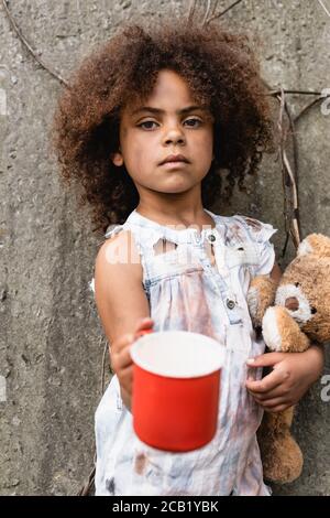 Fuoco selettivo del bambino africano americano povero che tiene l'orso del teddy e la tazza di metallo per le elemosine sulla strada urbana Foto Stock