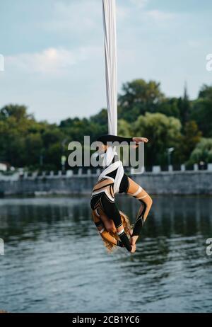 Baby girl aerialist esegue acrobazie su seta aerea appesa sullo sfondo di fiume, cielo e alberi. Foto Stock