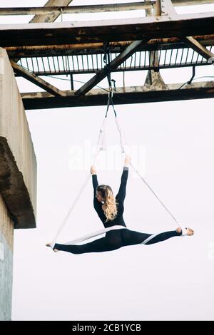 Donna aeralista esegue ginnastica split su seta aerea appesa attaccata al supporto del ponte sullo sfondo del cielo. Foto Stock