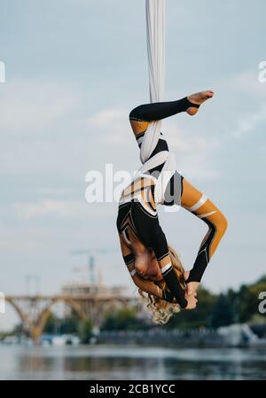 Baby girl aerialist esegue acrobazie su seta aerea appesa sullo sfondo di fiume, cielo e ponte. Foto Stock